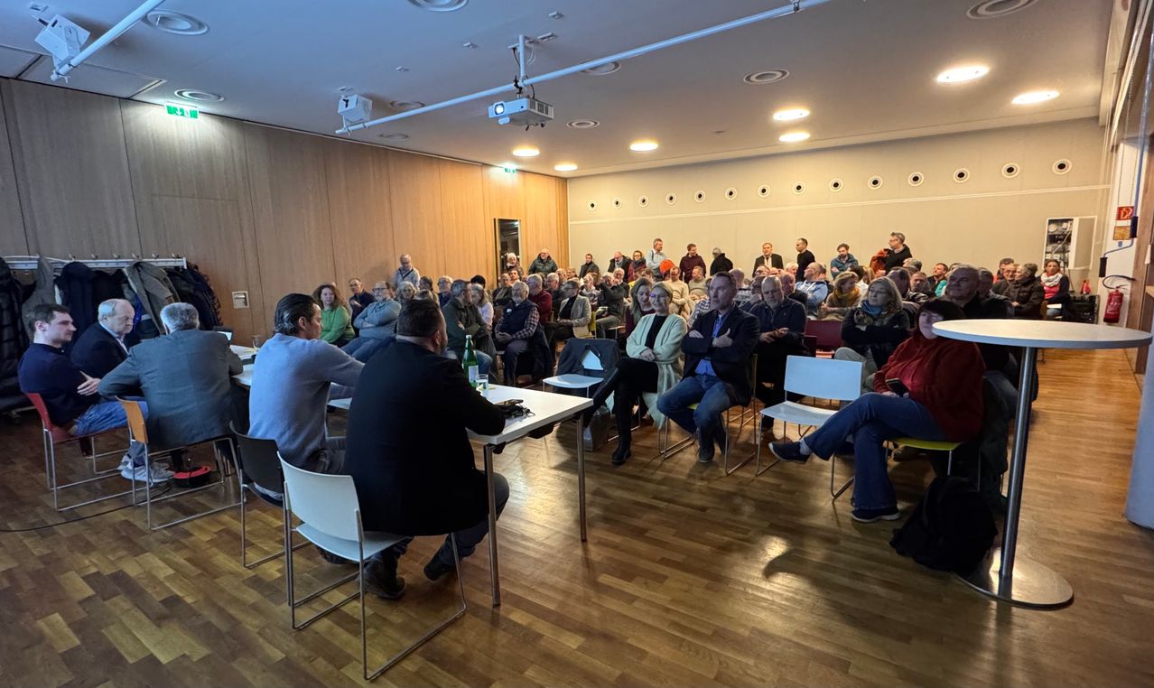 Ein Konferenzsaal mit Holzboden und -wänden, gefüllt mit Menschen, die auf Stühlen sitzen, einige stehend. Zwei Männer sitzen an einem Tisch davor, möglicherweise sprechend. Projektoren und Lampen hängen von der Decke.