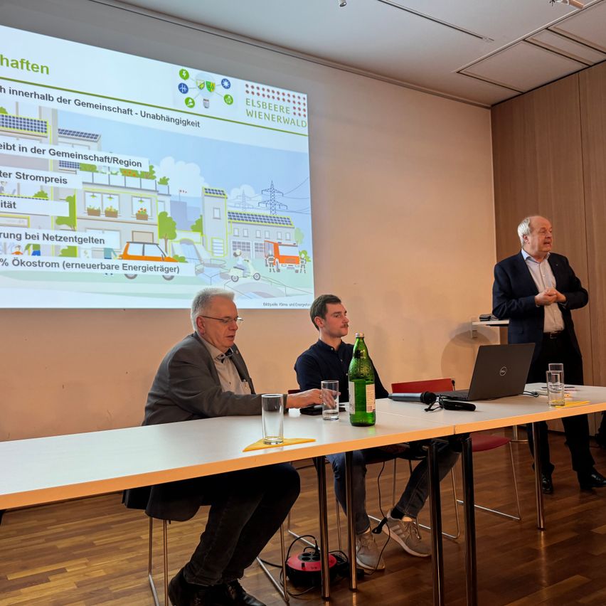 Drei Männer sitzen an einem Tisch mit einem Laptop, einem Mikrofon und einer Flasche davor. Eine Projektionsleinwand zeigt ein Diagramm. Ein Mann steht hinter ihnen an einem Podium.