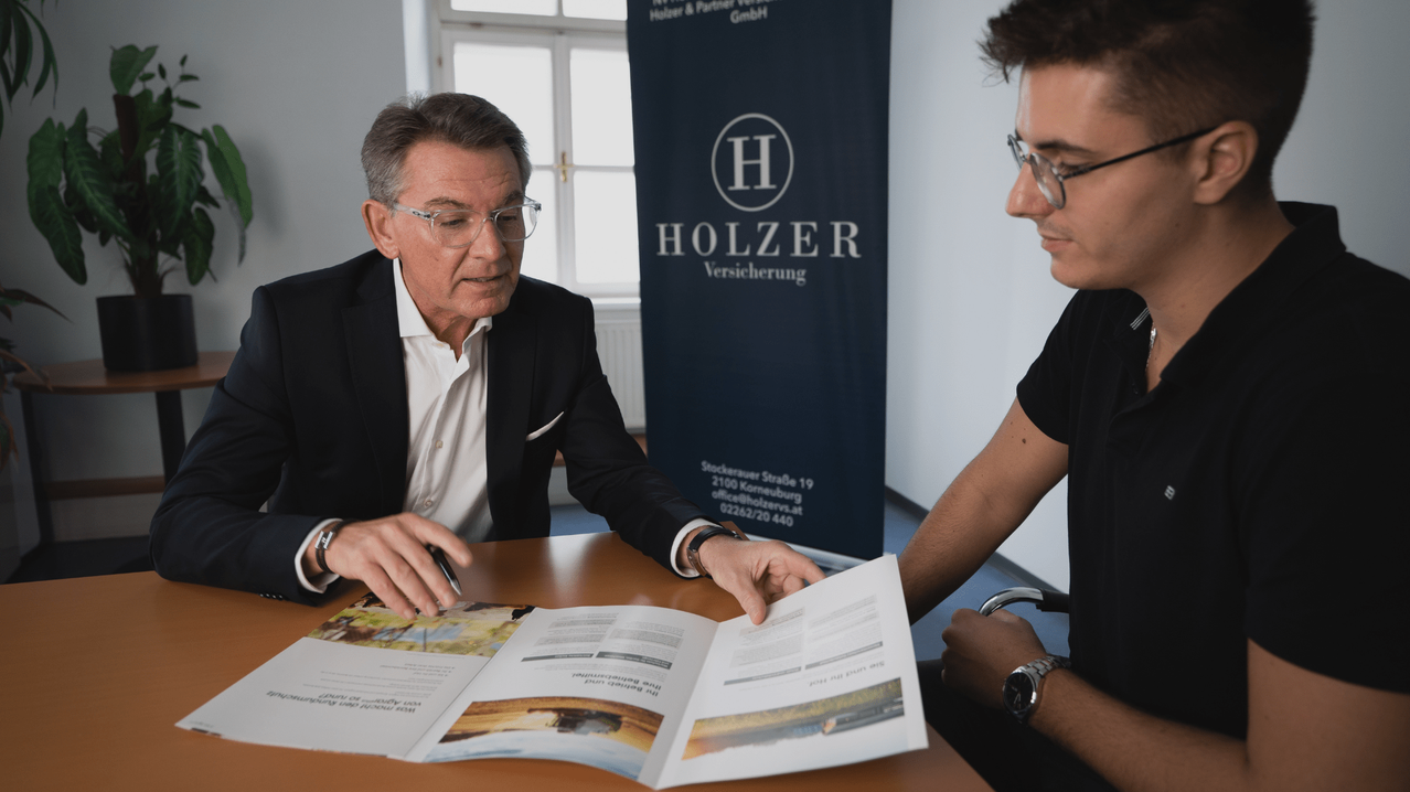 Ein Mann in Anzug weist mit seinem Stift auf einen Prospekt. Ein junger Mann in einem schwarzen T-Shirt sieht zu.