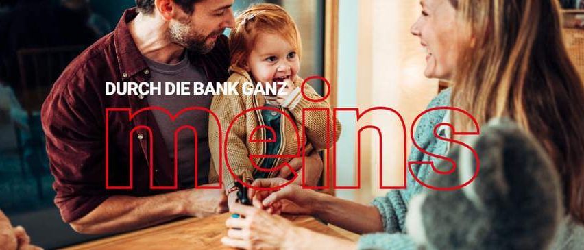 Eine Familie lächelt sich um einen Tisch herum an. Der Text lautet Durch die Bank ganz meins.