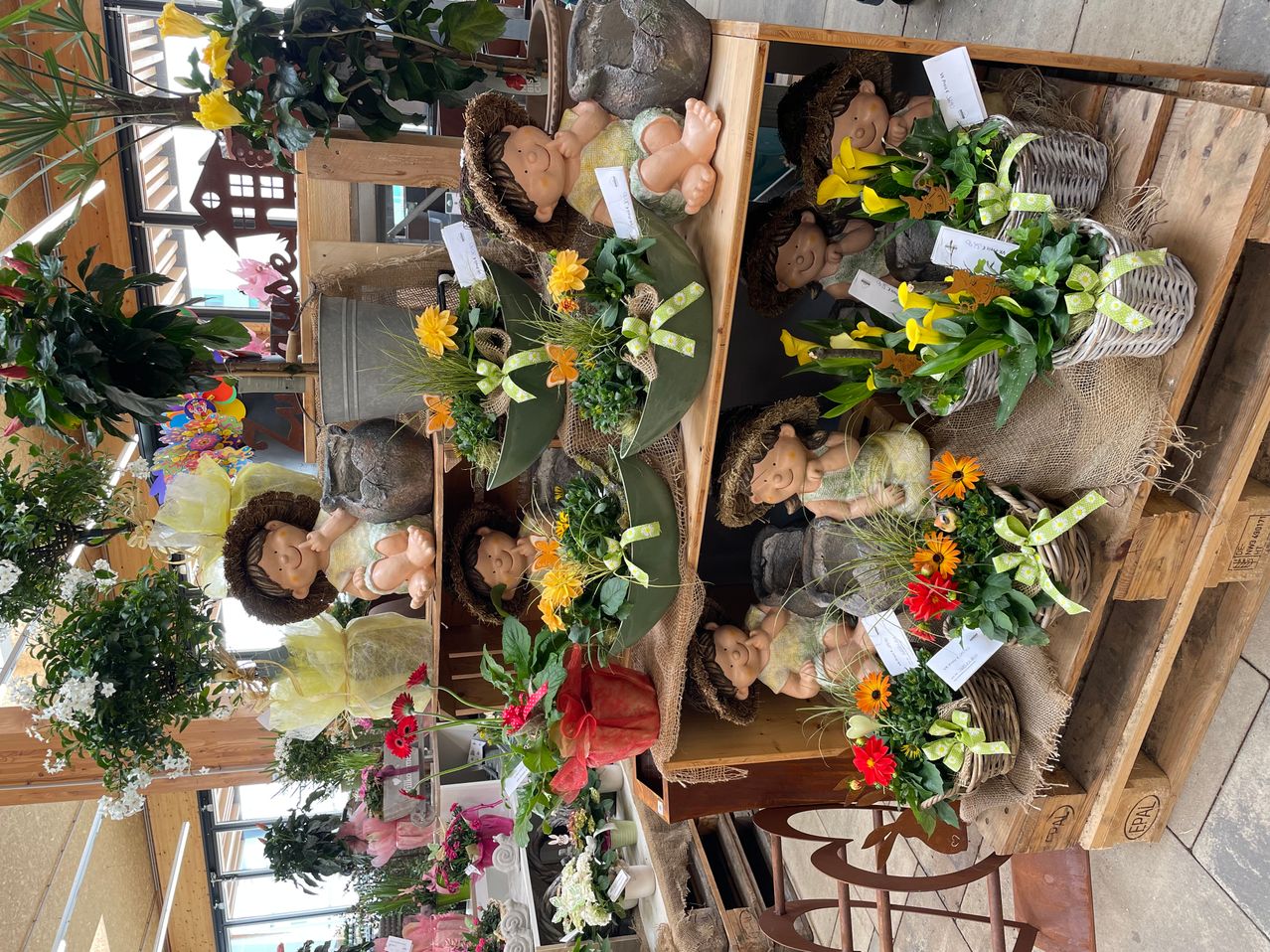 Drei Holzregale mit Körben, die mit Blumen und Babyfiguren gefüllt sind. Jeder Korb hat eine andere Blumenanordnung mit Bändern und Karten.