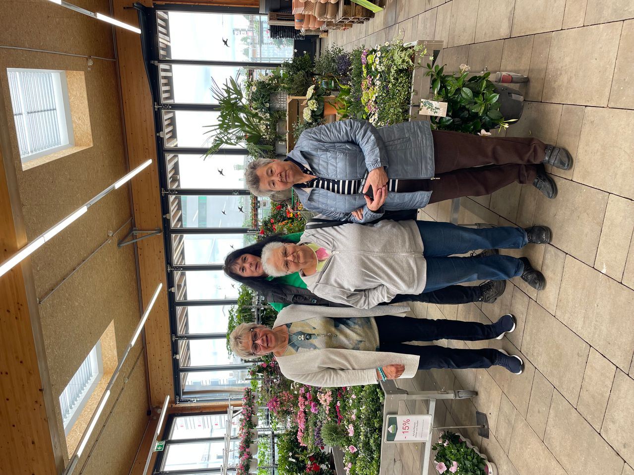 Drei Frauen stehen in einem Blumenladen und lächeln für ein Foto. Sie sind von verschiedenen Blumen und Pflanzen umgeben.