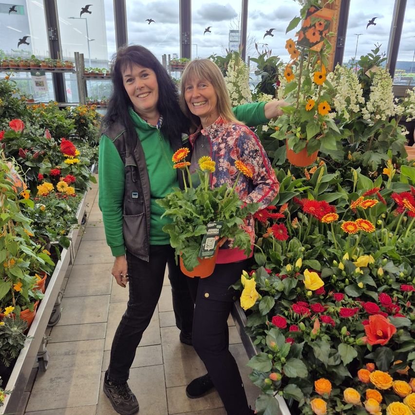 Zwei Frauen stehen in einem Blumenladen, lächeln und halten eine Topfpflanze mit orangen und gelben Blumen.