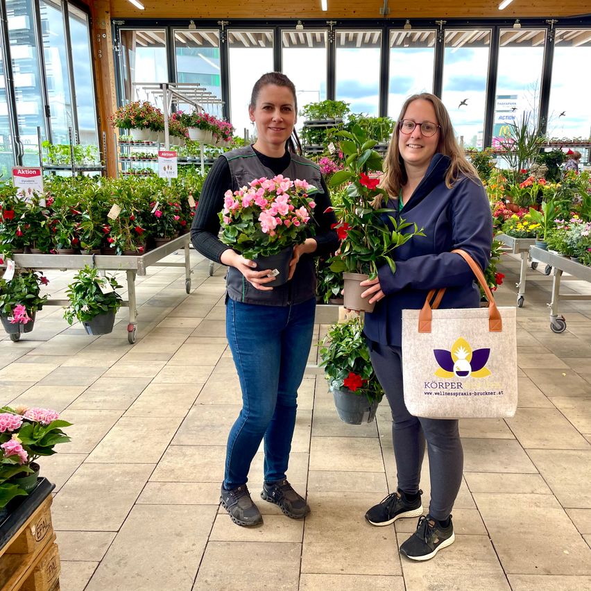 Zwei Frauen stehen in einem Pflanzenladen und halten eine Topfpflanze und eine Tasche. Eine Frau hat rosa Blumen und die andere Frau hat eine Tasche mit Logo. Hinter ihnen stehen viele Topfpflanzen auf Wagen.