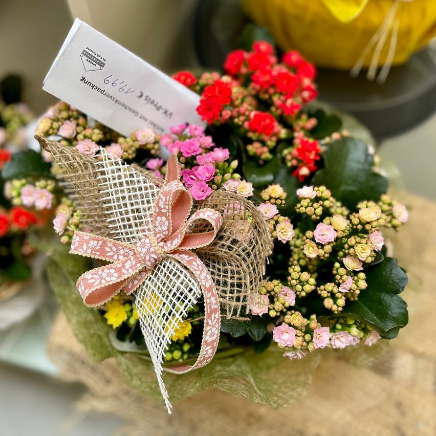 Eine Nahaufnahme eines Blumenarrangements mit einer rosa Schleife, einem Schild mit Text und einer Vielzahl von Blumen in Rot, Rosa und Gelb.