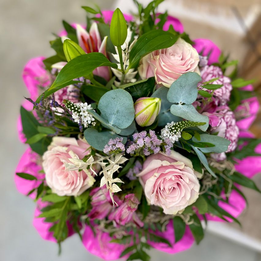 Ein Nahaufnahme eines Blumenarrangements zeigt einen Strauß aus rosa Rosen, Flieder und grünen Blättern. Die Rosen sind kreisförmig angeordnet, mit einigen Blättern verstreut. Die Blumen sind in voller Blüte, und das Arrangement ist ordentlich angeordnet.