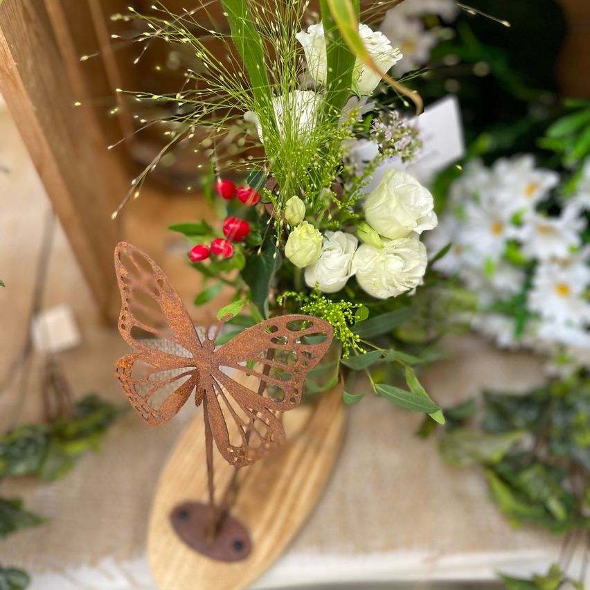 Eine dekorative Blumenarrangement mit weißen Rosen, roten Beeren und grünen Blättern, auf einem Holzsockel mit einer rostigen Schmetterlingsmotiv präsentiert.