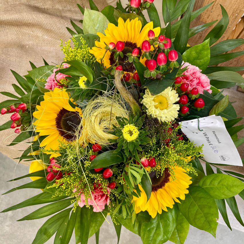 Ein lebendiges Blumenarrangement besteht aus einer Mischung von Sonnenblumen, Gänseblümchen, rosa Blumen und roten Beeren, verziert mit grünen Blättern und einem weißen Etikett.