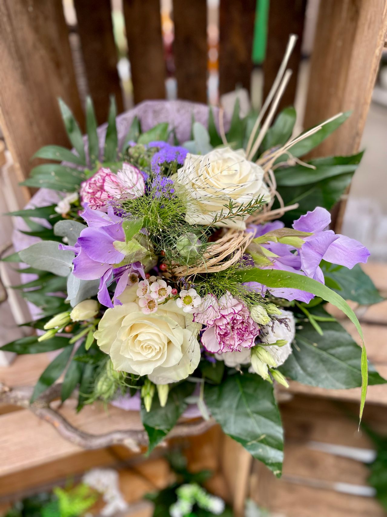Ein Blumenarrangement mit weißen Rosen, lila Blumen und grünen Blättern. Der Strauß ist auf einer Holzoberfläche arrangiert.