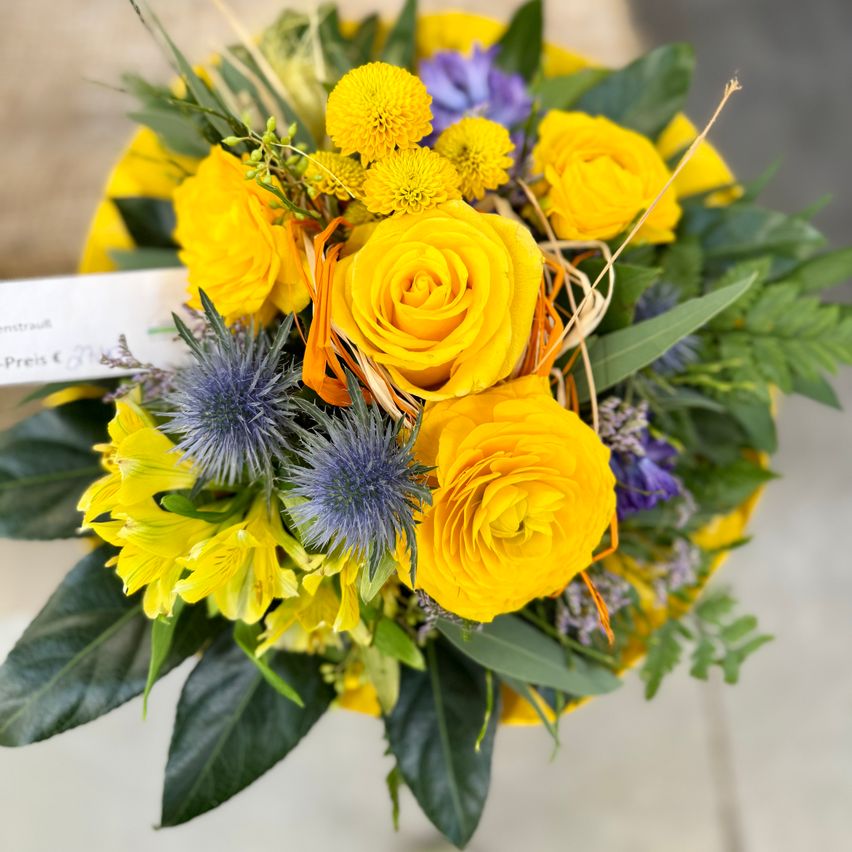 Ein gelber Blumenstrauß mit leuchtend gelben Rosen, lila Blumen und grünen Blättern, präsentiert in einem gelben Korb mit Preisschild.