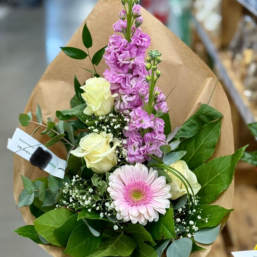 Ein Blumenstrauß in braunem Geschenkpapier enthält pinke und weiße Rosen, lila Delphinien und hellpinke Gerbera-Gänseblümchen, umgeben von grünen Blättern.