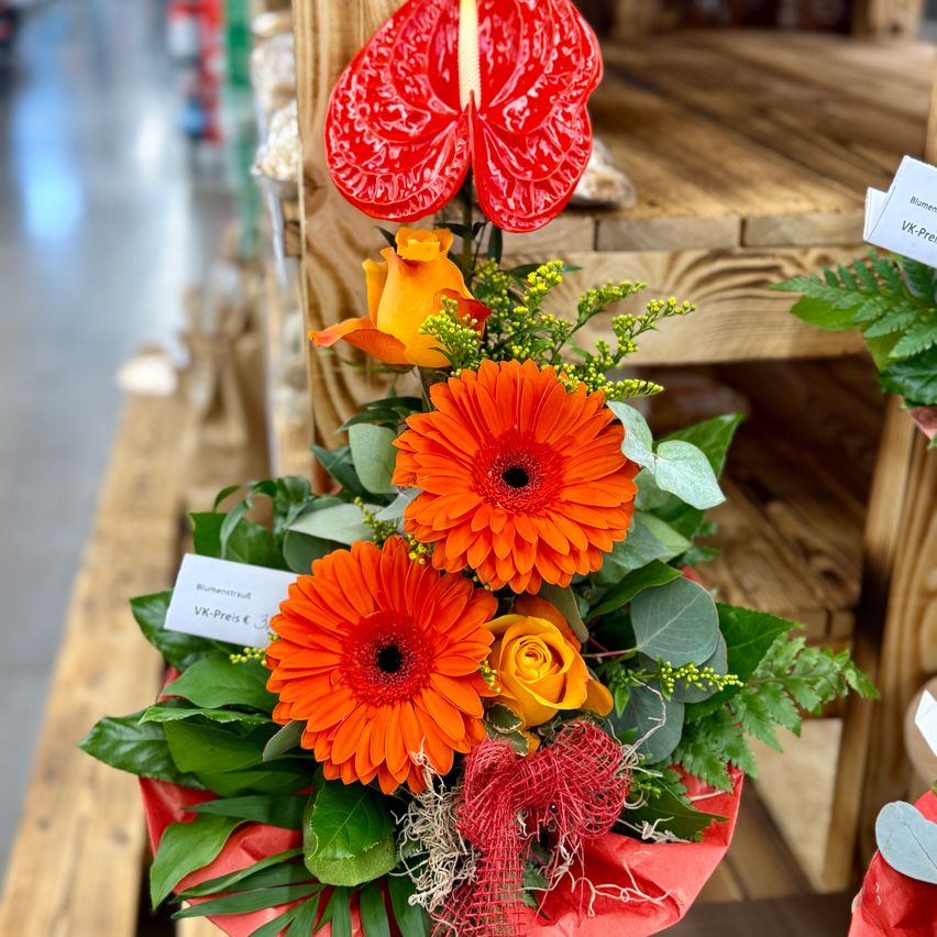Ein leuchtender Strauß mit roten und orangefarbenen Blumen, einschließlich Rosen und Gänseblümchen, in einem roten Behälter mit Grünpflanzen und einem roten Band. Der Strauß wird in einem Geschäft mit Holzregalen ausgestellt.