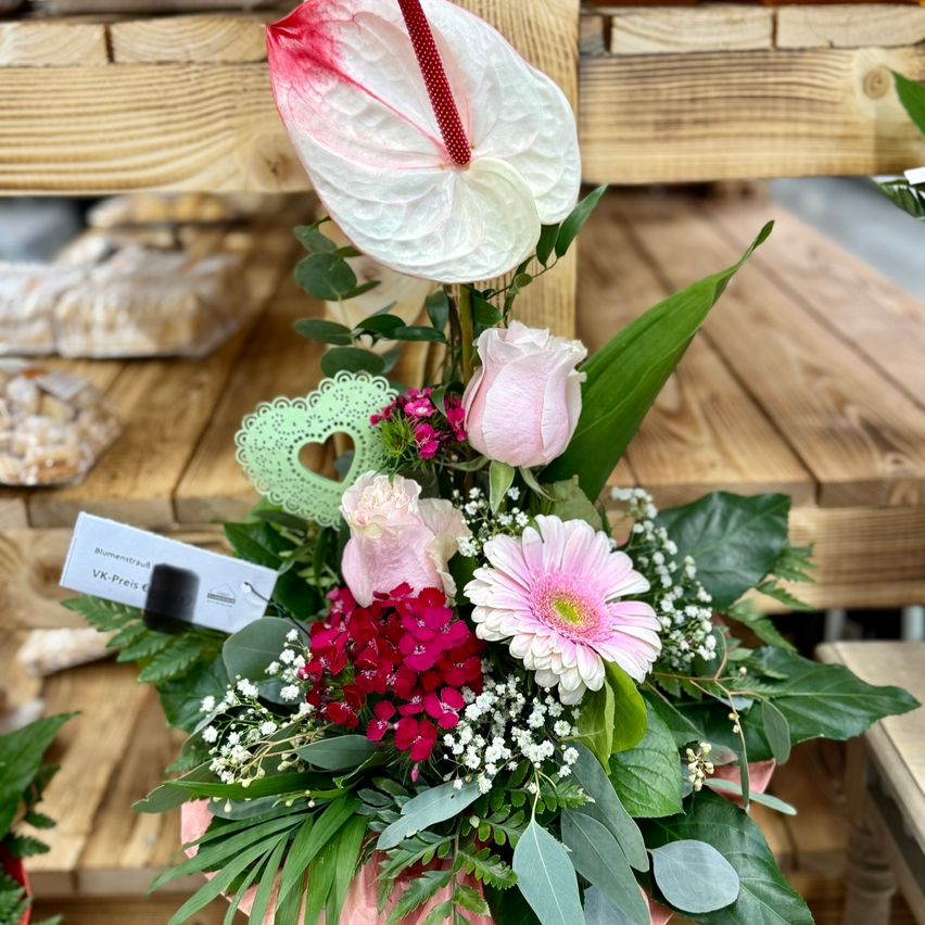 Eine pinke Blumenarrangement mit weißen und rosa Rosen, einer rosa Margerite und einer roten Blume auf einem Holzregal.