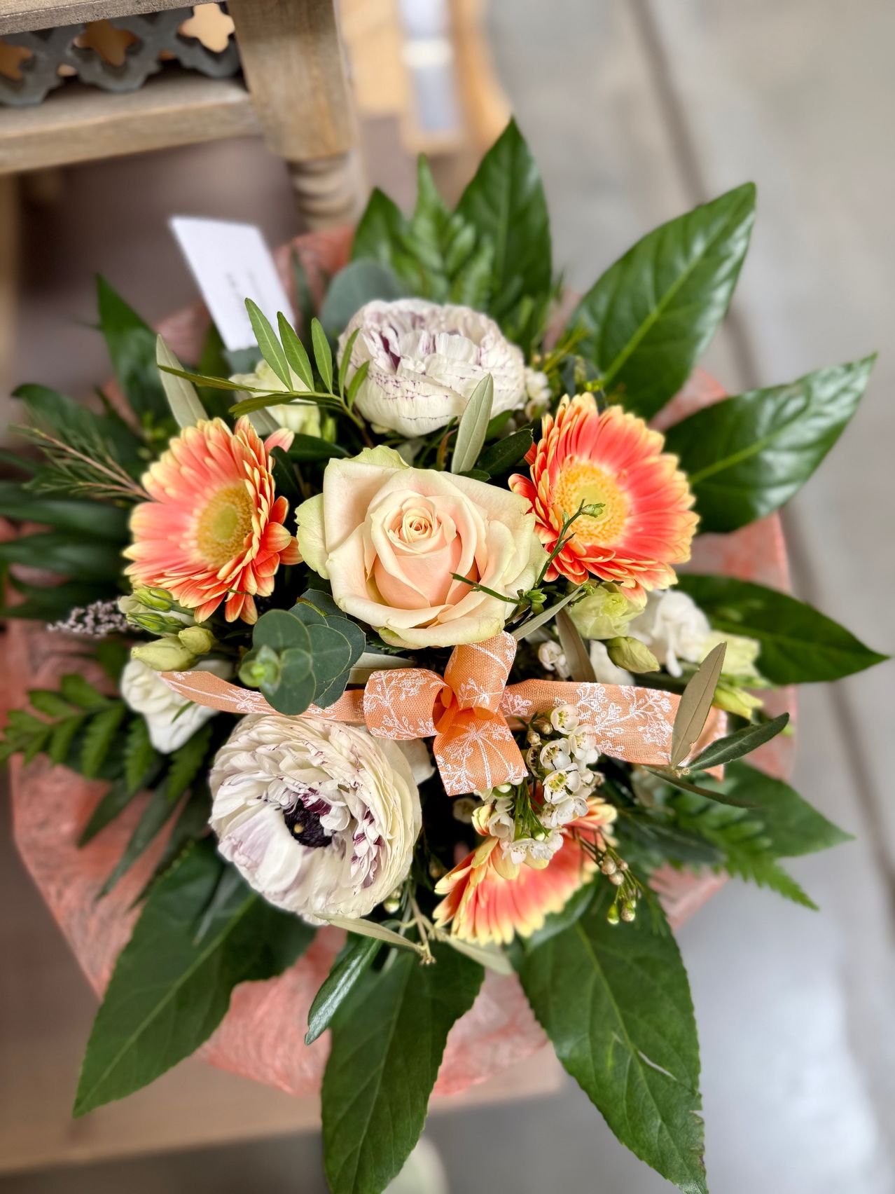 A bouquet of flowers with pink, white, and orange petals. The flowers are arranged in a circular shape, with green leaves surrounding them. The flowers have a ribbon tied around them.