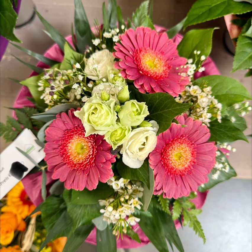 A close-up of a pink gerbera daisy bouquet with white and green flowers and leaves. The flowers have red centers and yellow petals.