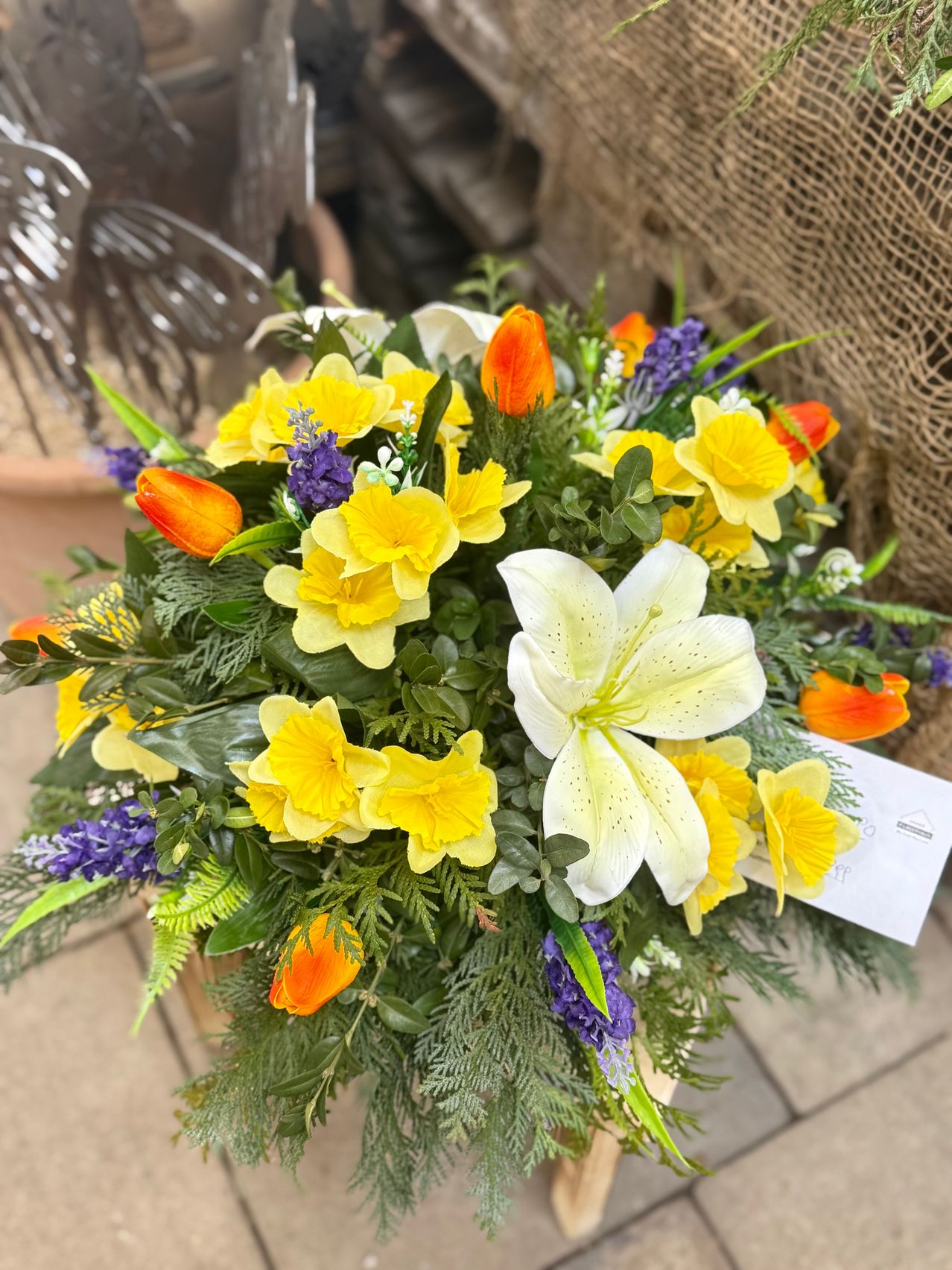 Ein Blumenarrangement zeigt gelbe und orange Narzissen, eine auffällige weiße Lilie und violette Veilchen. Grüne Blätter und Stängel umgeben die Blumen, mit einem sichtbaren Etikett im Vordergrund.