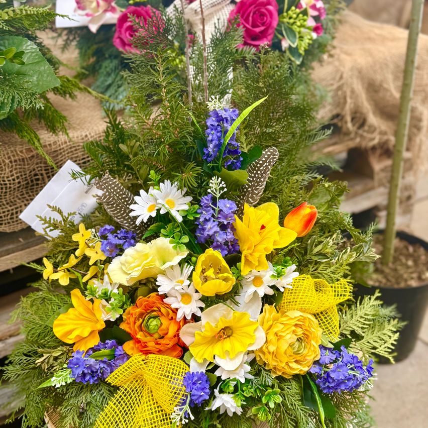 Eine lebhafte Blumenarrangement besteht aus einer Mischung von gelben, orangefarbenen und weißen Blumen, ergänzt durch blaue und lila Blüten, alles in einem Korb mit einem gelben Band arrangiert.