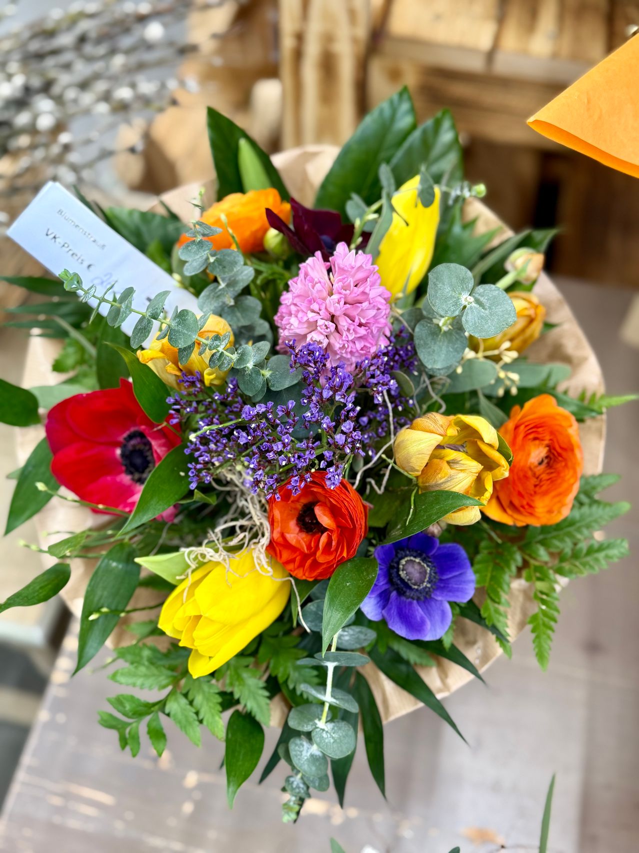 Ein farbenfroher Strauß mit einer Mischung aus roten, gelben, orangefarbenen und rosa Blüten, zusammen mit lila und grünem Laub, ist in braunes Papier eingewickelt. Ein weißer Karton ist am Strauß befestigt.