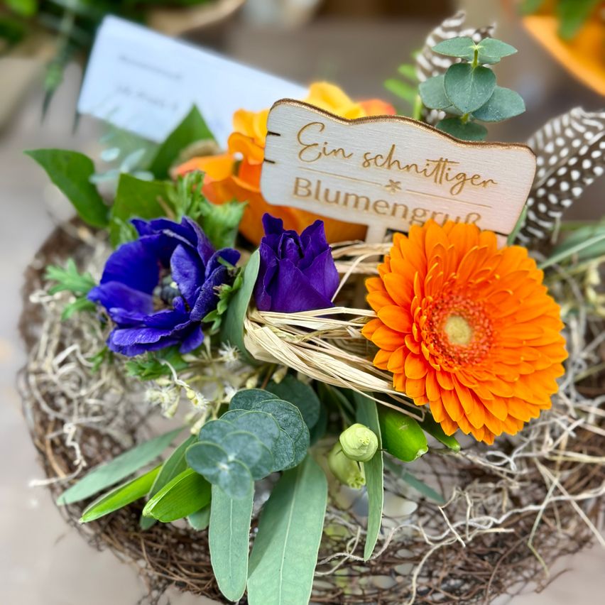 Ein Korb mit Blumen und einem Schild, das 'Ein schnittiger Blumengruß' steht. Die Blumen sind in Lila, Blau und Orange. Die Anordnung besteht aus verschiedenen grünen Blättern und Stroh.