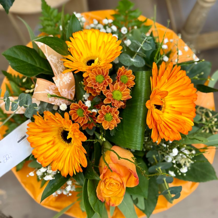 Ein farbenfroher Blumenstrauß mit orangen und gelben Gänseblümchen, einer orangen Rose und weißen Babys atem, umgeben von üppigen grünen Blättern, alles auf einer orangen Basis arrangiert.