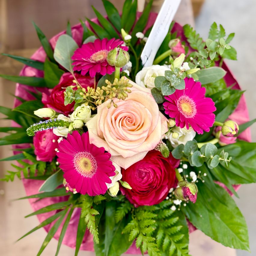 Ein Strauß aus rosa, weißen und roten Blumen, einschließlich Rosen und Gerberas, in einer rosa Verpackung mit grünen Blättern arrangiert.