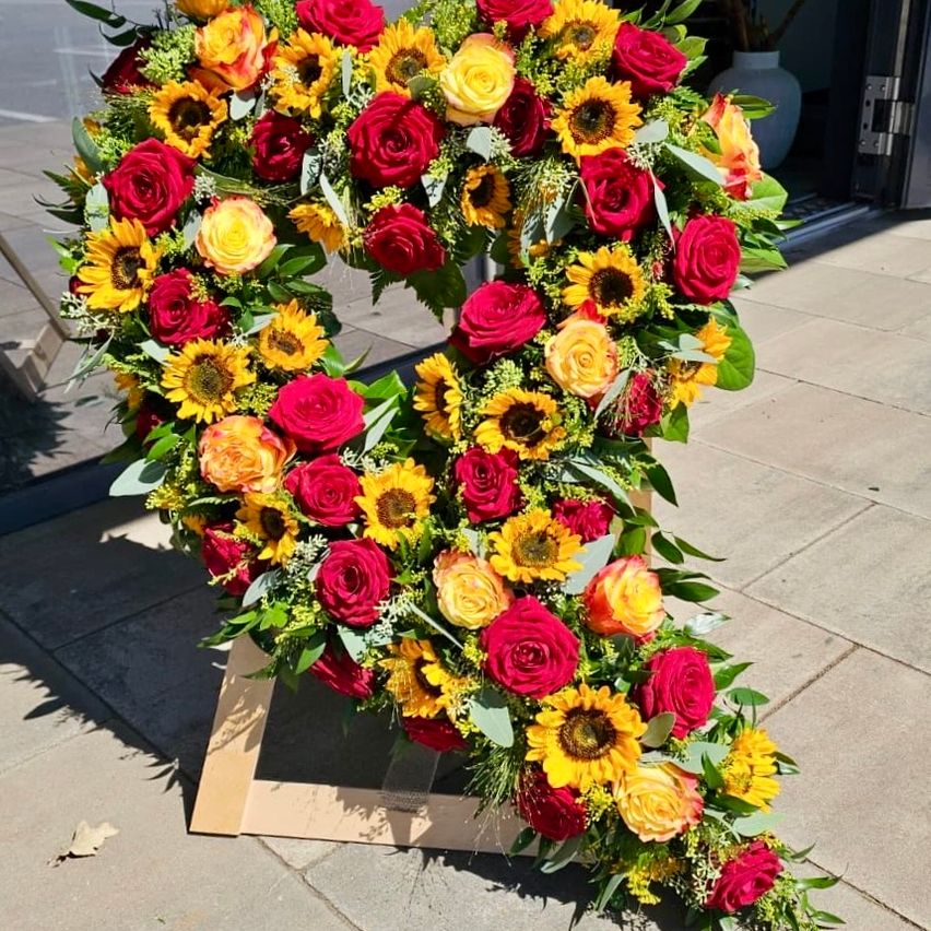 A heart-shaped floral arrangement with red roses, yellow sunflowers, and green leaves on a wooden stand on a sunny day.