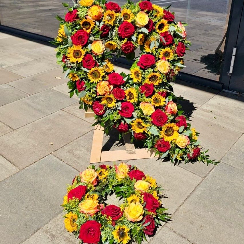 A floral arrangement shaped like the number 8, composed of red roses, yellow sunflowers, and green leaves, placed on a wooden stand on a paved surface.