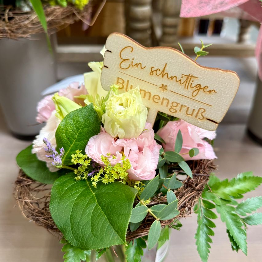 Ein Blumenarrangement mit rosa und gelben Blumen in einem Korb. Ein Holzschild trägt den Schriftzug 'Ein Schnittiger Blumengruß'. Die Blumen sind in voller Blüte.
