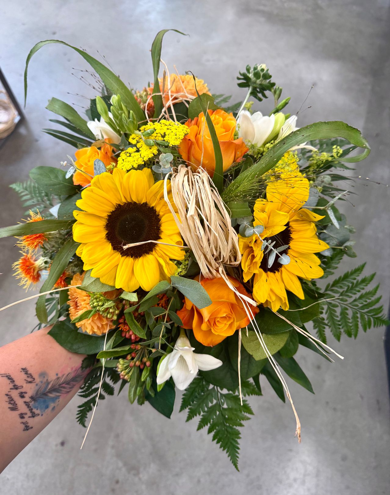 Ein leuchtender Strauß aus Sonnenblumen, orangefarbenen Rosen und grünem Laub. Der Strauß wird von jemandem gehalten, der eine Tätowierung am Arm hat.