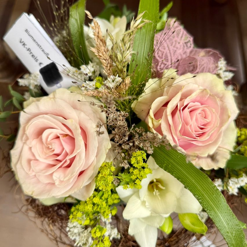 Ein Strauß aus rosa Rosen, weißen Lilien und grünen Blättern. Die Komposition beinhaltet einen Zettel mit Text.