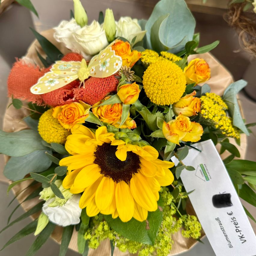 Ein Strauß mit gelben und weißen Blumen, einschließlich Rosen, Sonnenblumen und dekorativen Blättern, mit einem gelben Schmetterling darauf. Die Komposition steht auf einer braunen Basis und enthält einen weißen Anhänger.