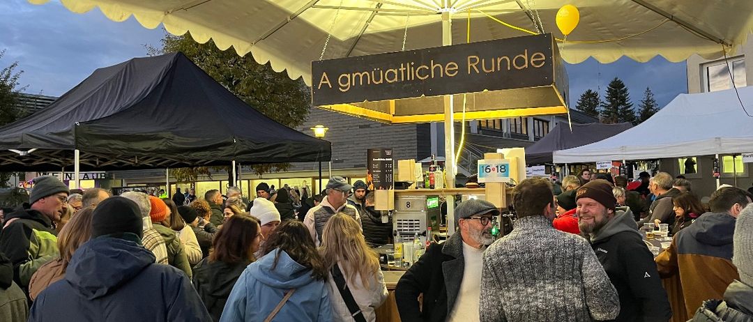 Eine Menschenmenge versammelt sich an einem Außenstand mit einem großen Schirm, der mit 'A gmuatlich Runde' beschriftet ist. Der Stand ist von Menschen umgeben, einige tragen Winterkleidung. Bäume und Gebäude sind im Hintergrund zu sehen.