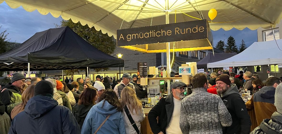 Eine Menschenmenge versammelt sich an einem Außenstand mit einem großen Schirm, der mit 'A gmuatlich Runde' beschriftet ist. Der Stand ist von Menschen umgeben, einige tragen Winterkleidung. Bäume und Gebäude sind im Hintergrund zu sehen.