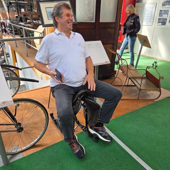 Ein Mann sitzt auf einem alten Fahrradsitz und lächelt, während er ein Handy in der Hand hält. Hinter ihm geht eine Frau. Die Szene spielt sich in einem Museum mit Fahrrädern und alten Möbeln ab.