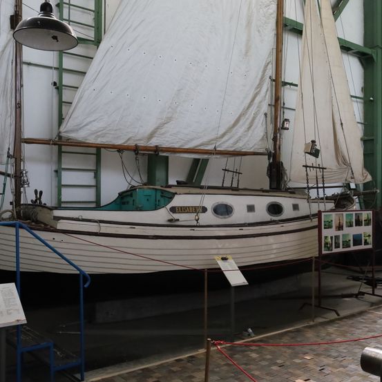 Ein Segelboot namens 'ELISABETH' wird in einem Museum ausgestellt, mit weißem Tuch bedeckt, mit Seilen und einer Leiter in der Nähe.