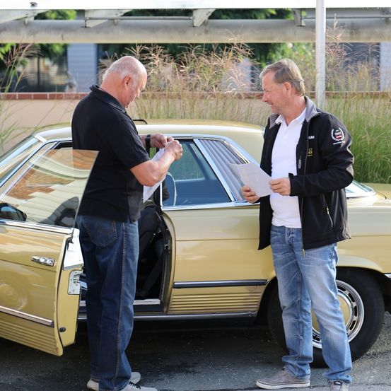 Zwei Männer stehen neben einem gelben Oldtimer, einer der Männer hält ein Papier und der andere schaut lächelnd darauf.