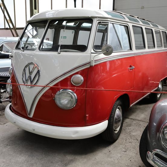 Ein klassischer roter und weißer Volkswagen Typ 2 Bus steht in einer Garage mit anderen Autos, darunter ein silberner.
