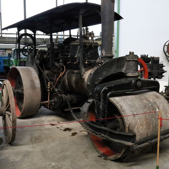 Ein alter dampfbetriebener Straßenwalze wird in einer Garage mit Betonboden ausgestellt. Der Walze hat große Räder und eine Dampfmaschine.