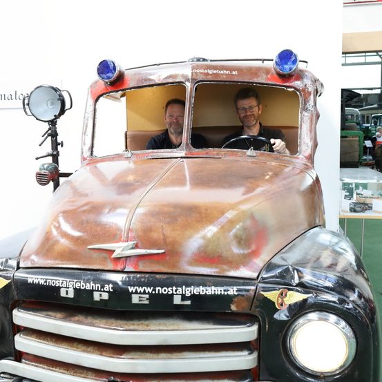 Zwei Männer sitzen in einem alten Opel-LKW und lächeln. Vorne steht 'OPEL' und 'www.nostalgiebahn.at' darunter. Oben sind Lichter und links eine Lampe.