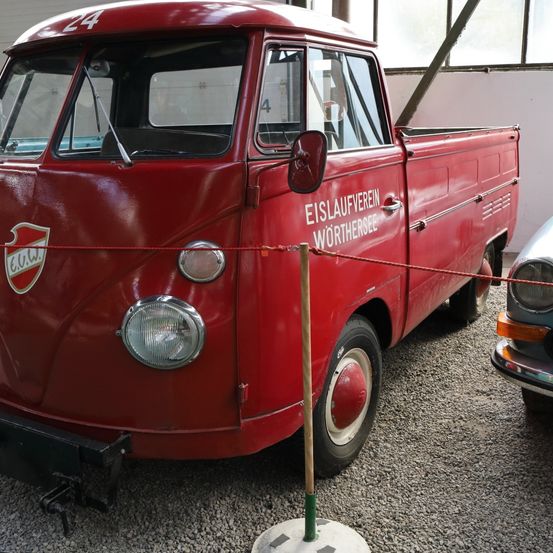 Ein roter Vintage-Pickup-Truck mit der Nummer 24 wird in einer Garage ausgestellt. Der Truck hat einen roten Schild auf der Front und den Text "EISLAUFVEREIN WÖRTHERSEE" an der Seite.