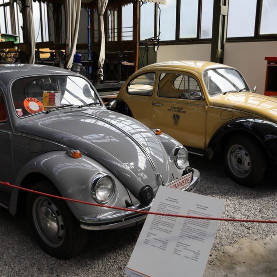 Zwei Vintage-Volkswagen-Käfer, einer silber und einer gelb, sind in einer Garage geparkt mit Informationsschildern. Der gelbe hat ein rotes Licht oben.