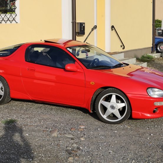 Ein roter Sportwagen steht vor einem gelben Gebäude mit Stufen und einer Geländer. Der Wagen hat silberne Felgen und eine glänzende Karosserie. Ein blaues Auto ist im Hintergrund teilweise zu sehen.