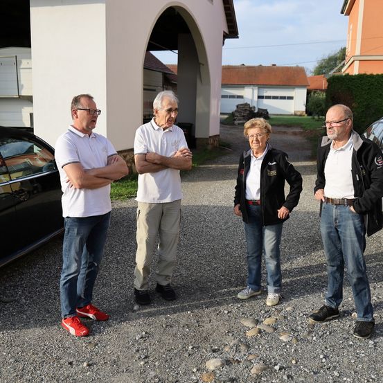 Vier ältere Personen, zwei Männer und zwei Frauen, stehen in einem Schotterbereich vor einem Gebäude. Sie scheinen ein Gespräch zu führen.
