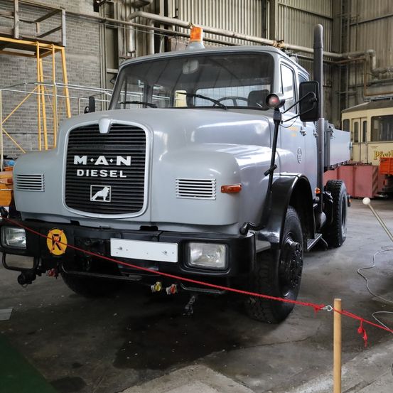 Ein grauer MAN Diesel LKW ist in einer Lagerhalle geparkt, vor ihm befindet sich eine rote Absperrung. Hinter dem LKW steht eine Ziegelmauer und ein gelbes Gerüst.