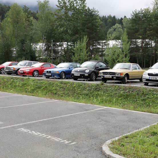Mehrere Autos sind auf einem Parkplatz geparkt, mit Bäumen im Hintergrund. Ein Auto hat das Wort 'Reserviert' auf dem Boden geschrieben.