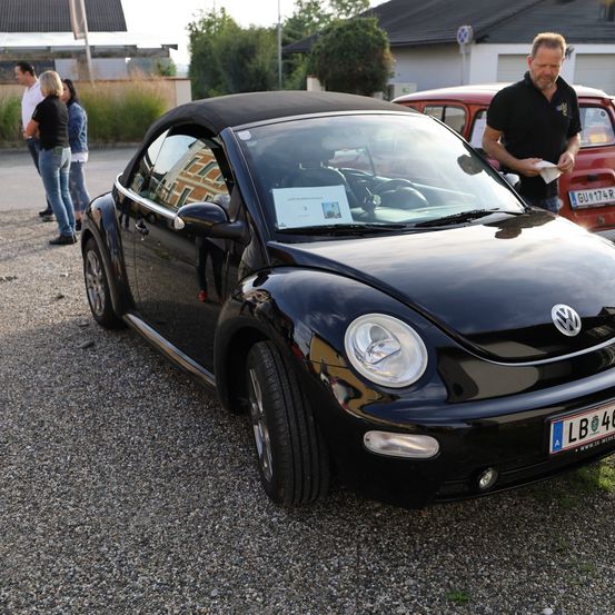 Ein schwarzer Cabrio-Volkswagen Käfer ist auf Kies mit dem Nummernschild LB 40 geparkt. Ein Mann liest davor eine Zeitung, während andere dahinter stehen.