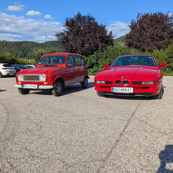 Zwei rote Autos sind auf einem Parkplatz geparkt. Eines ist ein Renault und das andere ein BMW. Im Hintergrund befinden sich Bäume und Büsche, und in der Ferne Berge.