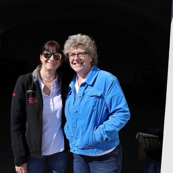 Zwei Frauen stehen lächelnd für die Kamera in einem Türrahmen. Eine trägt eine schwarze Jacke, die andere eine blaue Jeansjacke. Beide haben eine Brille auf und sind in lässiger Kleidung gekleidet.