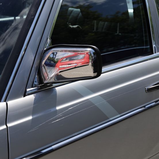 Nahaufnahme eines seitlichen Autospiegels in Silber, der ein rotes Auto und Bäume reflektiert. Das Auto hat silberne Verzierungen und eine kleine Delle.
