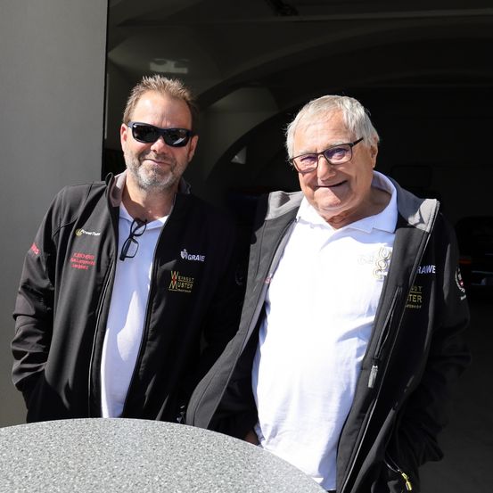 Zwei Männer stehen vor einer Garage, beide tragen Sonnenbrillen und Jacken. Ein Mann lächelt, während der andere zur Seite schaut. Der Mann auf der rechten Seite hat eine Brille und ein weißes Hemd.